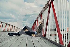 An attractive young man with long blonde hair, in berets and a military coat is leaning on a pyril bridge