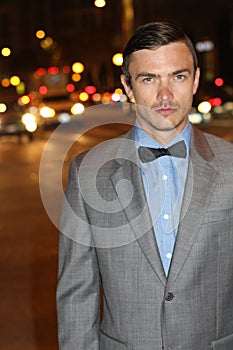 Attractive young man at night with city lights behind him, wearing elegant suit jacket and bow tie