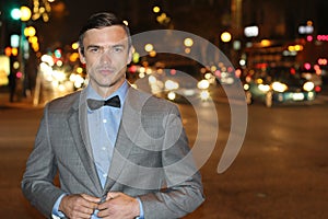 Attractive young man at night with city lights behind him, wearing elegant suit jacket and bow tie with copy space