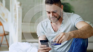 Attractive young man with smartphone and credit card shopping on the internet sit on bed at home