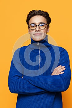 an attractive young man stands on an orange background in a blue zip-up jacket and black eyeglasses and looks into the