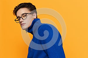 an attractive young man stands on an orange background in a blue zip-up jacket and black eyeglasses and looks into the