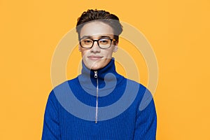 an attractive young man stands on an orange background in a blue zip-up jacket and black eyeglasses and looks into the
