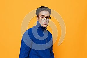 an attractive young man stands on an orange background in a blue zip-up jacket and black eyeglasses and looks into the