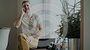 Attractive young man talking phone sitting on windowsill with laptop and camera at home
