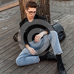 Attractive young man tourist in fashionable jeans wear in glasses with leather black backpack is resting near tree on street.