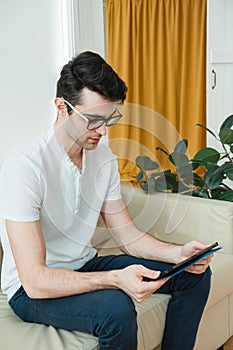 Attractive young man is using a tablet while sitting on the couch at home and browsing social networks and news.
