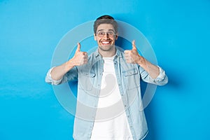 Attractive young man wearing glasses and casual clothes, showing thumbs up in approval, like something, standing against