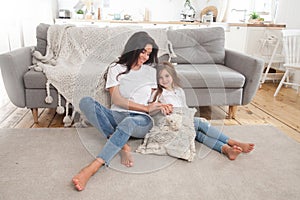 Attractive young mom with little cute girl are spending time together at home while sitting on the floor by the sofa with grey