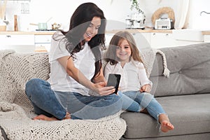 Attractive young mom and little daughter playing online games at home using smartphone, having video chat or watching movie.