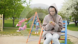 Attractive young mother typing message on smartphone while her children ride on swing in park