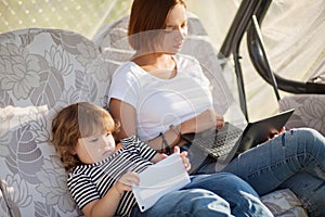 Attractive young mother using laptop, while daughter playing tablet.