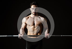 Attractive young muscular athlete posing with a barbell on a dark background