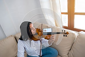 An attractive young musician plays the violin sitting on the sofa. The girl is practicing playing a musical instrument at home