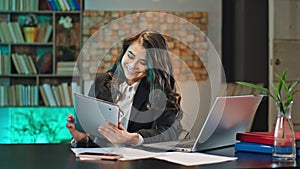 Attractive young office manager woman at her work place working on the electronic tablet and laptop at the same time