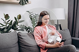 Attractive young pregnant woman applying headphones on belly sitting alone on comfy couch at home. Happy expectant mother