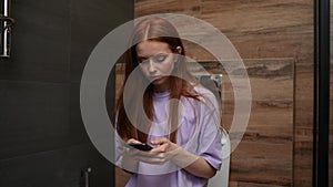 Attractive young redhead woman using smartphone sitting on toilet bowl at home bathroom with modern interior.