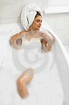 Attractive young relaxed brunette woman wearing white towel on her head enjoying her bath with bubbles in bright bathroom