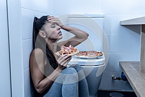 Attractive young and sad bulimic young woman feeling guilty and sick eating while sitting on the floor next to the toilet in