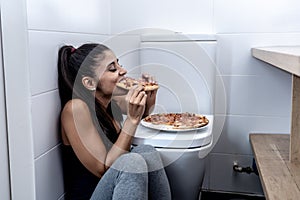Attractive young and sad bulimic young woman feeling guilty and sick eating while sitting on the floor next to the toilet in