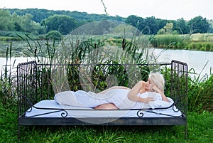 Attractive, young, sexy, blonde woman lying naked covered with white duvet in bed sleeping in the nature in front of a lake