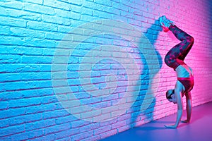 Attractive young slim gymnast woman in sports clothing stretching on brick wall in neon lights. Flexible muscular woman