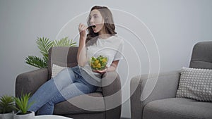 Attractive young slim woman holding big bowl with salad enjoying her healthy food while sitting in the arm-chair