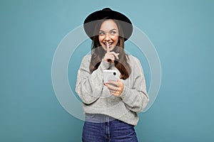 Attractive young smiling woman wearing black hat and grey sweater holding smartphone looking to the side showing shhh