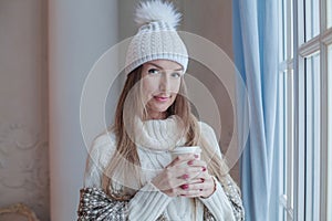 Attractive young smiling woman in white woolen sweater and knitted hat at cozy home