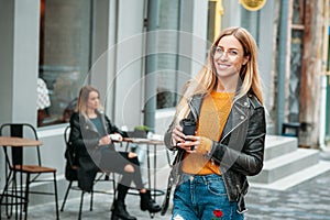 attractive young stylish woman holding a cup of coffee and standing near cafe.