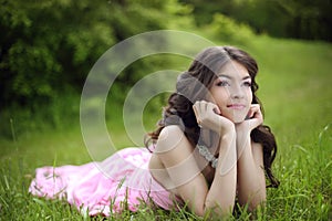 Attractive young teen girl with makeup wearing in pink dress lying on green grass field at spring park.