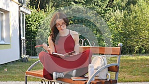 Attractive young tired girl in eyeglasses sitting on the brench outdoors, learning and drinking from termo cup some