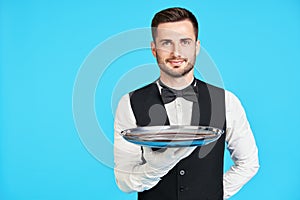Attractive young waiter holding empty silver tray over blue background