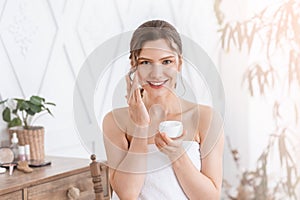 Attractive young woman applying face cream at home