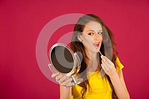 Attractive young woman applying lip gloss looking in mirror over pink background