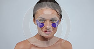 Attractive young woman applying medical eye patches under eyes and smiles looking at camera, isolated over gray