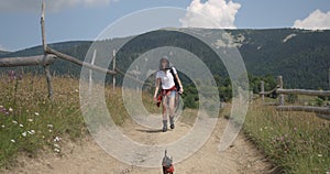 The attractive young woman with backpack is hiking with her dog and walking along the trail in beautiful mountains