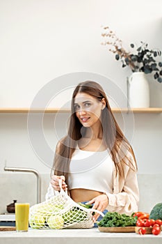 An attractive young woman with a bag full of fresh vegetables and fruits is standing in the kitchen