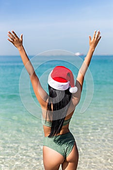 Attractive young woman in bikini with red christmas santa claus hat on ocean beach with hands up. Rear view