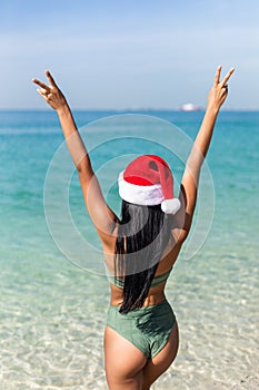 Attractive young woman in bikini with red christmas santa claus hat on ocean beach with hands up. Rear view