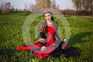 An attractive, young woman in a black and red medieval dress with a crown on her head sits in a field with a sword in her hands. C