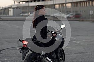 Attractive young woman in black tight-fitting suit and full-face protective helmet rides on sports motorcycle at urban