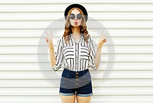 Attractive young woman blowing red lips sending sweet air kiss in black round hat, shorts, white striped shirt on white wall