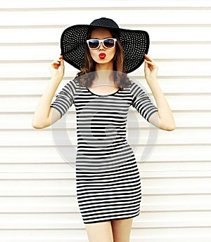 Attractive young woman blowing red lips sending sweet air kiss in black summer straw hat on white wall