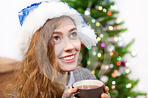 Attractive young woman in blue Santa hat holding hot tea mug in hands