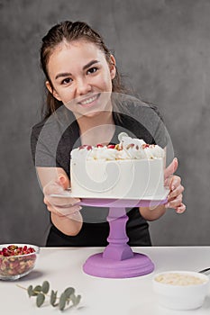 Attractive young woman brunette confectioner presents a white cake with small red flowers of a food rose.