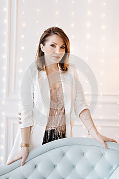 Attractive young woman in a business suit with white jacket, black pants and high heels near a blue classic sofa. White background