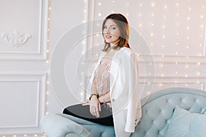 Attractive young woman in a business suit with white jacket playfully crouched on the side of a blue classic sofa. White