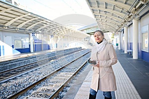 Attractive young woman commuter in beige coat, waiting to board the train on the railway station. Railroad trip