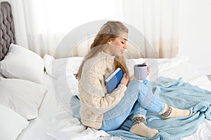 Attractive young woman in cozy warm sweater with cup of hot drink and book sitting on bed